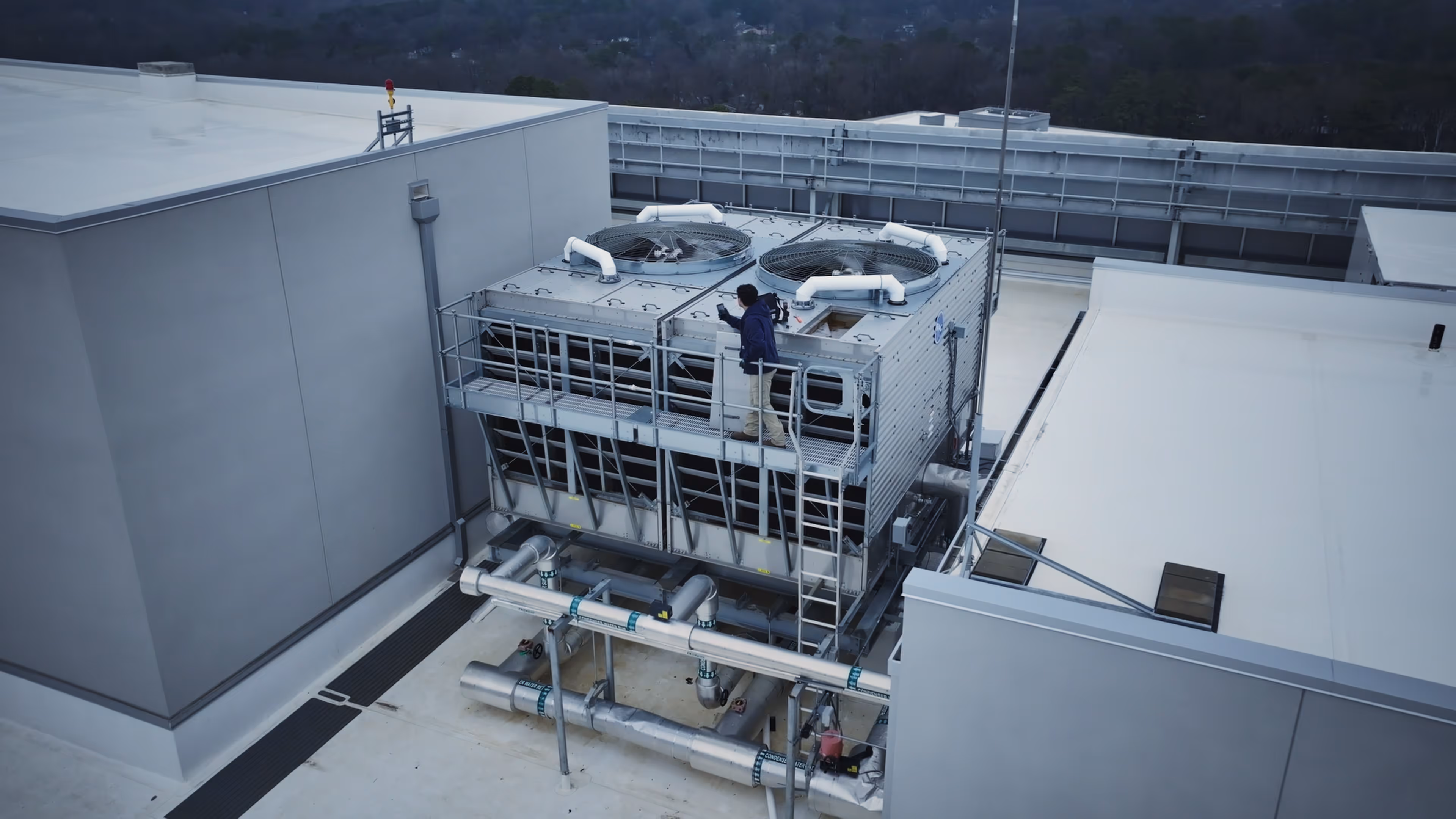 Technician inspecting large rooftop HVAC cooling unit with pipes and fans on a commercial building.