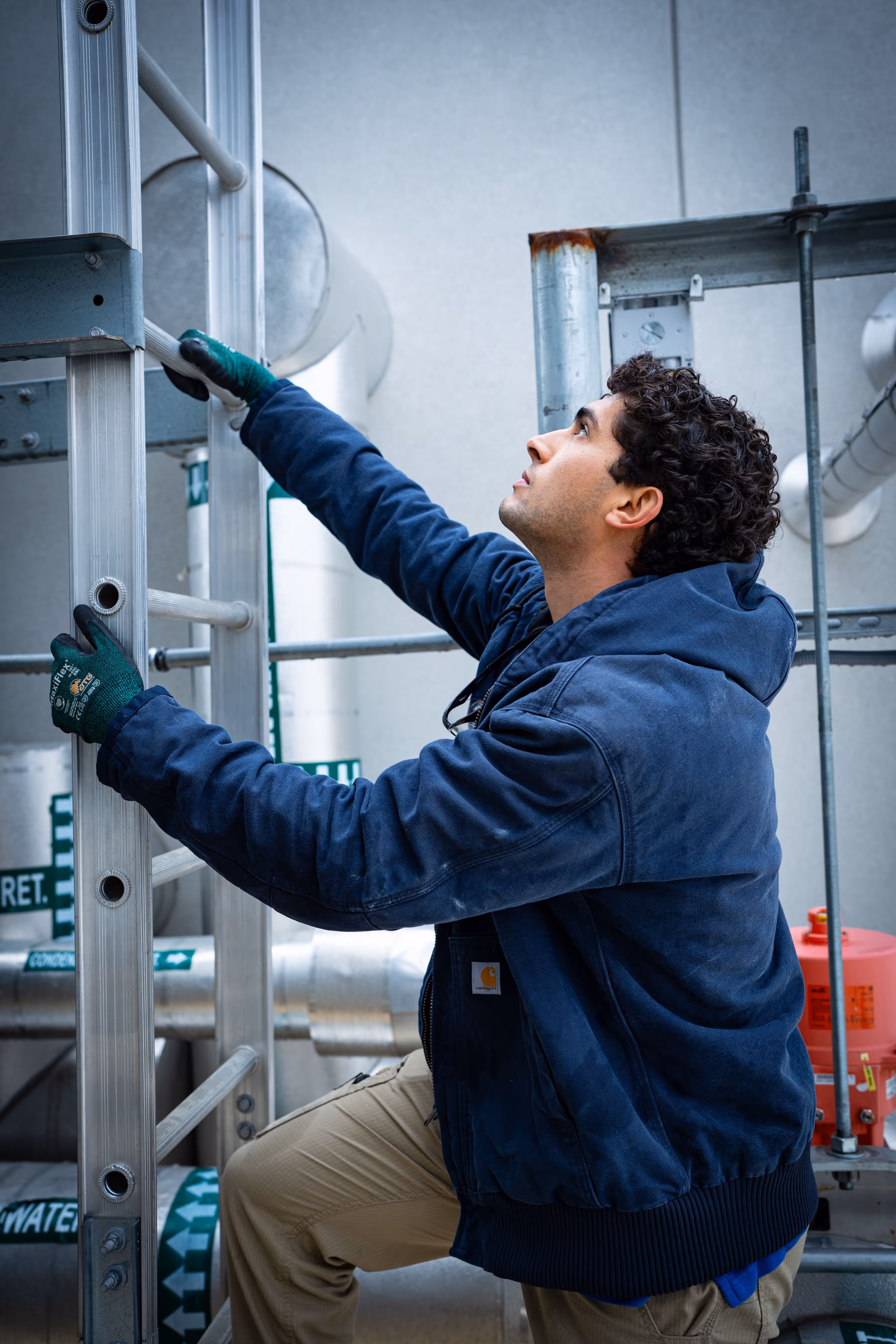 Man in blue jacket and gloves climbing a metal ladder in an industrial setting.