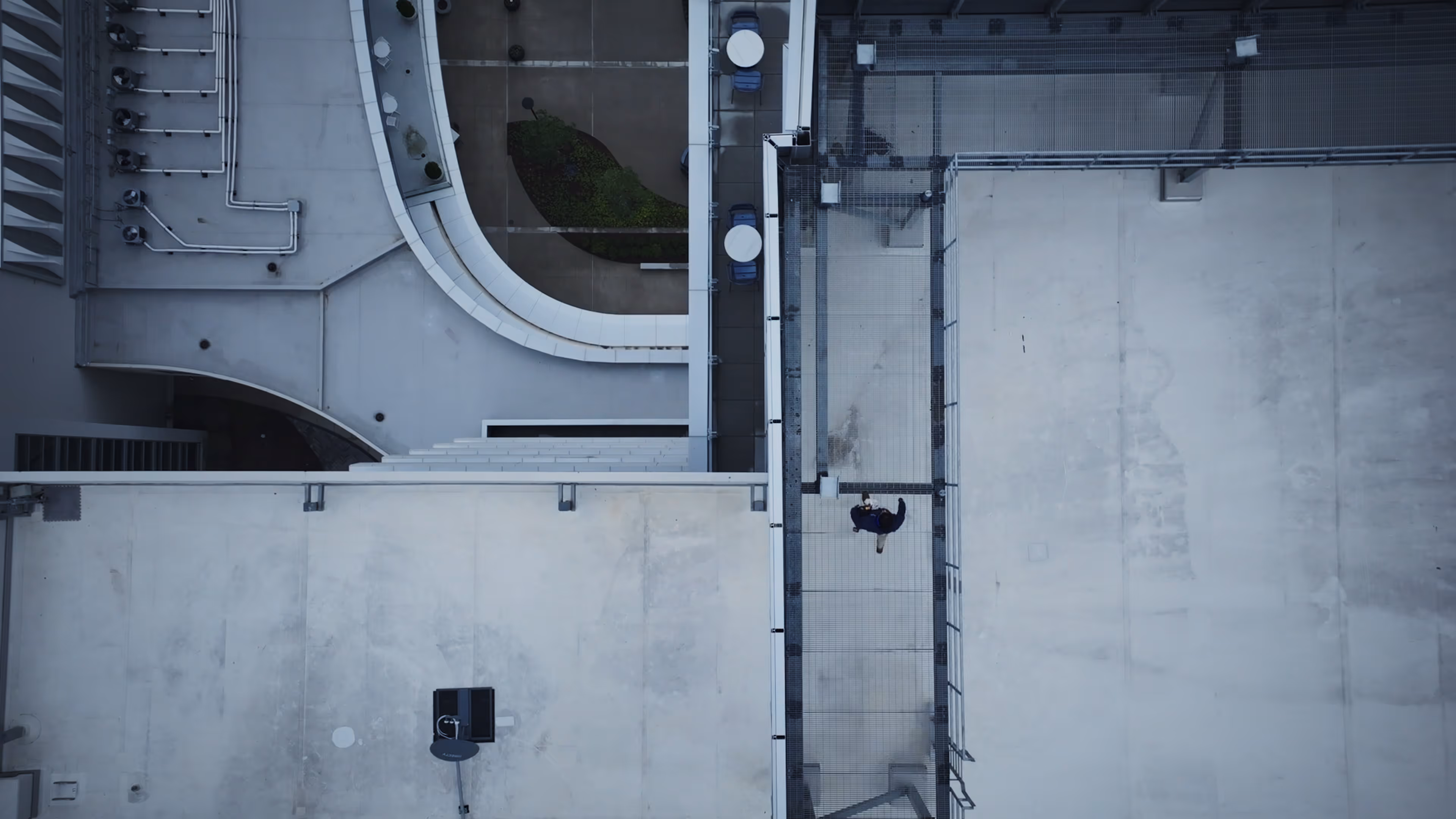 A person walking on a narrow rooftop walkway between two tall buildings, viewed from above.