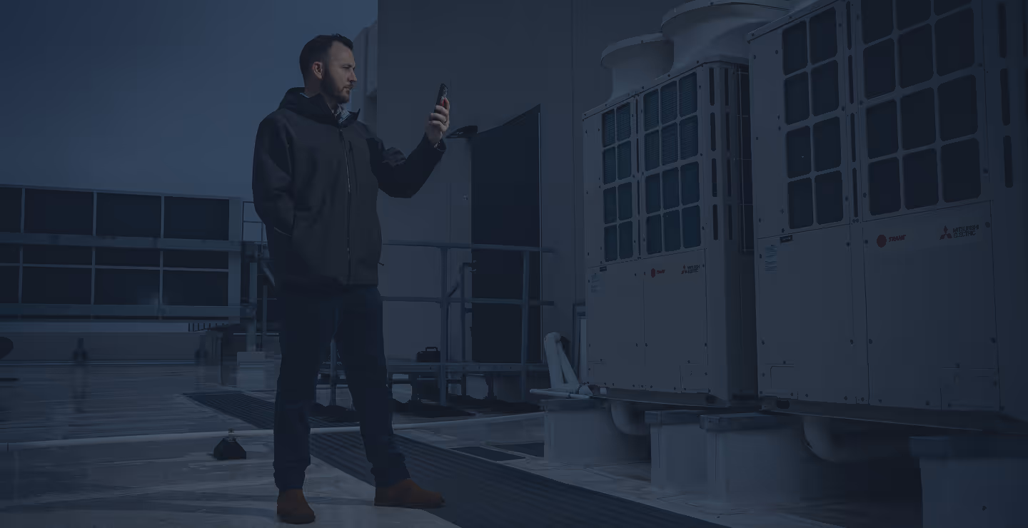 Man in black jacket using smartphone while inspecting large HVAC units on rooftop.