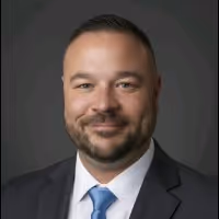 Smiling man with short hair and beard, wearing dark suit and blue tie against gray background.
