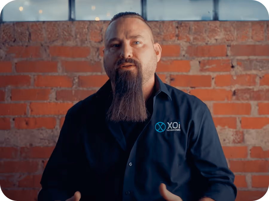 Man with a long beard wearing a black XOi Technologies shirt against a brick wall background.