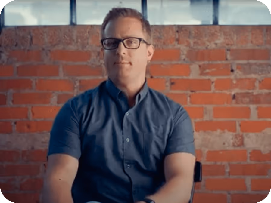 Man wearing glasses and a blue button-up shirt sitting in front of a red brick wall.