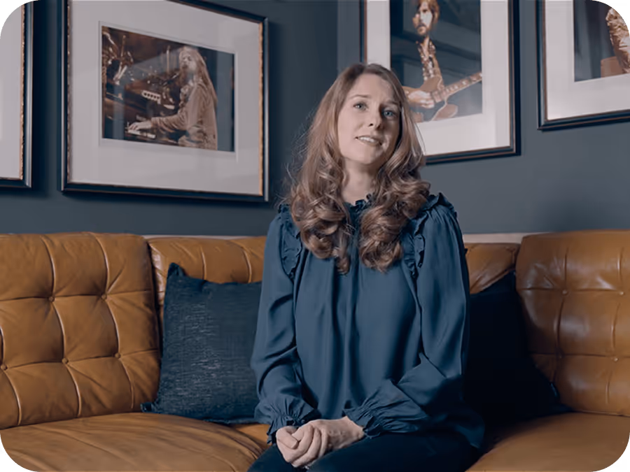 Woman with long curly hair sitting on a brown leather couch with music-themed photos on the wall.
