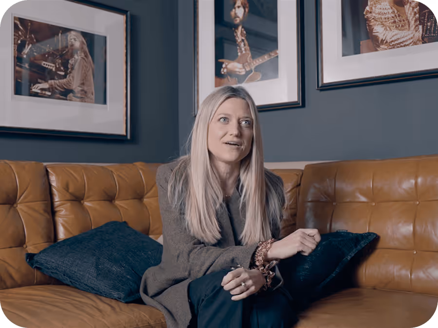 Blonde woman sitting on a brown leather couch with blue pillows, framed musician photos on dark wall.