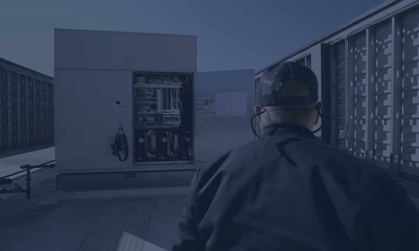 Technician in cap inspects open electrical panel of rooftop HVAC system outside.