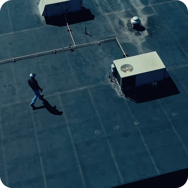 Person wearing a cap walking on a flat rooftop with ventilation units and pipes casting shadows.