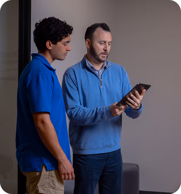 Two men in blue shirts standing and looking at a tablet together indoors.