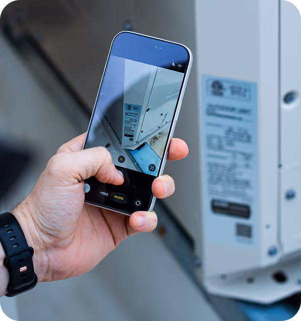 Hand holding smartphone taking a close-up photo of a white air conditioning unit's label.