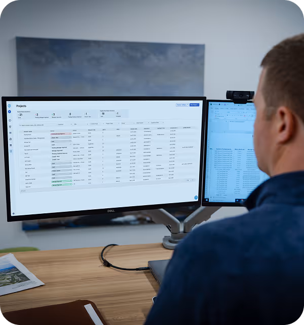 Person viewing project management data on dual computer monitors at a wooden desk.