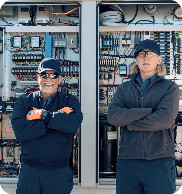 Two electrical technicians with arms crossed standing in front of open control panels with wiring.