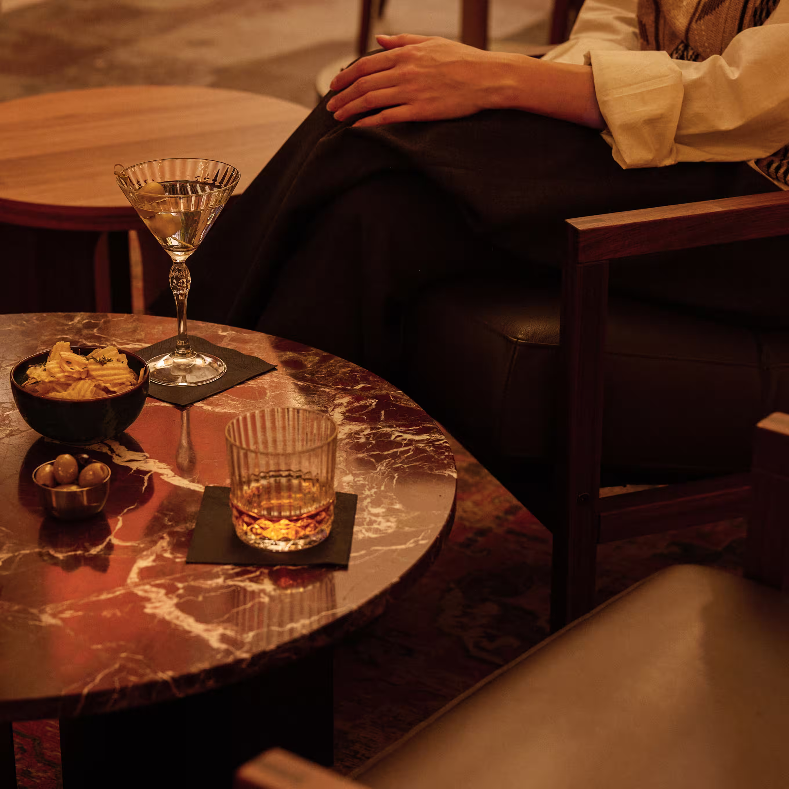 Cocktailhetkiä Taulu-baarissa – martini ja viskilasi sipsien kera punaisella marmoripöydällä, rentoilu nahkatuolissa