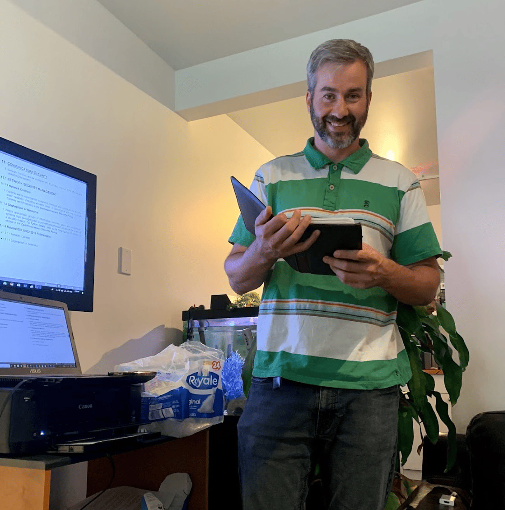 Man in green striped shirt smiling while holding an open binder in a room with computer monitors.