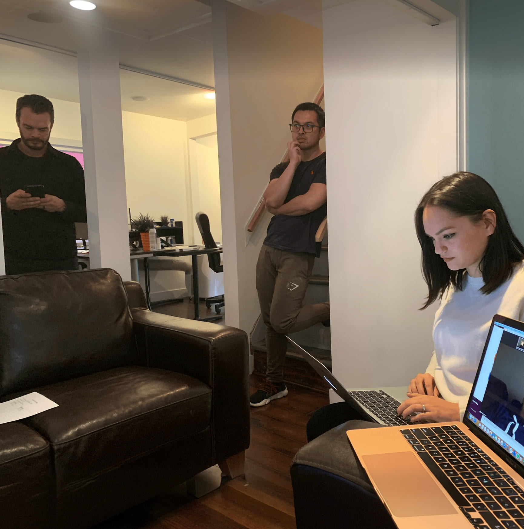 Three people in an office: one woman working on a laptop, two men standing and looking thoughtful.