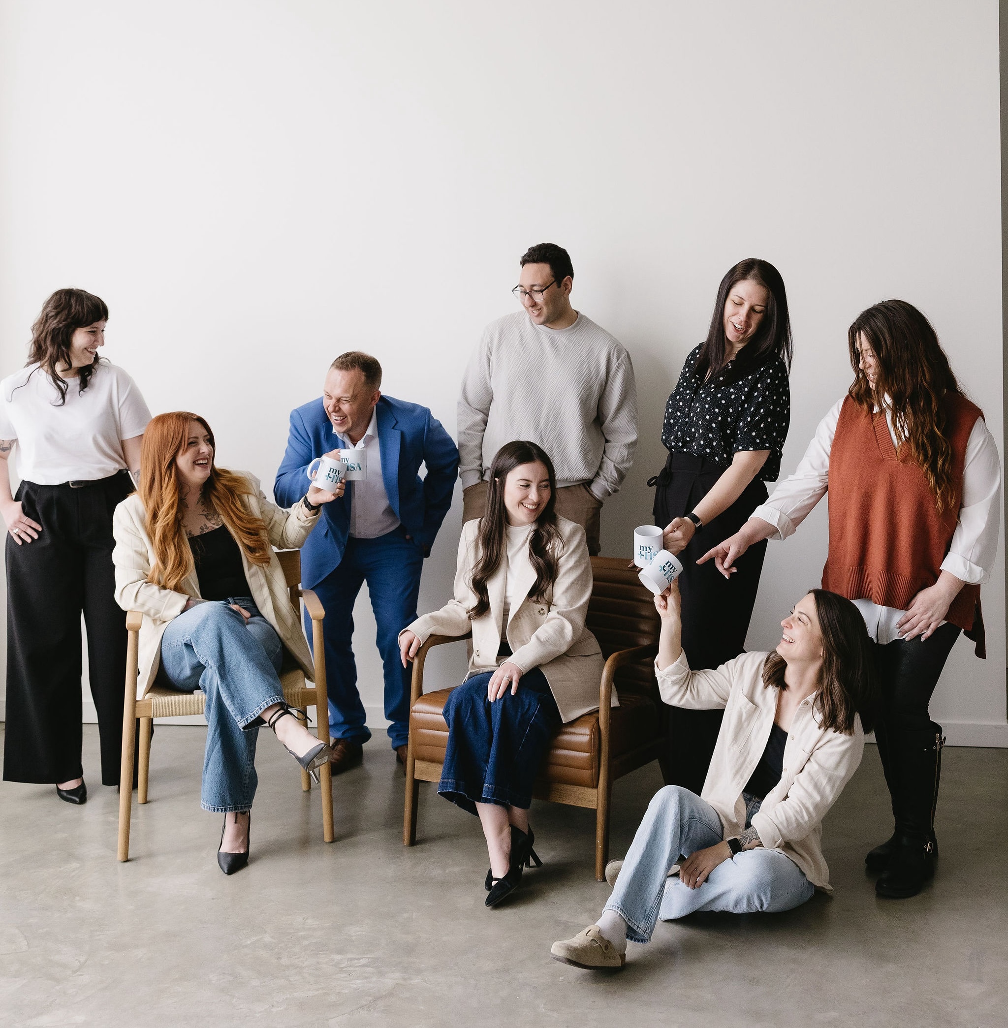 Group of seven people smiling and interacting, some seated, holding white mugs in a bright room.