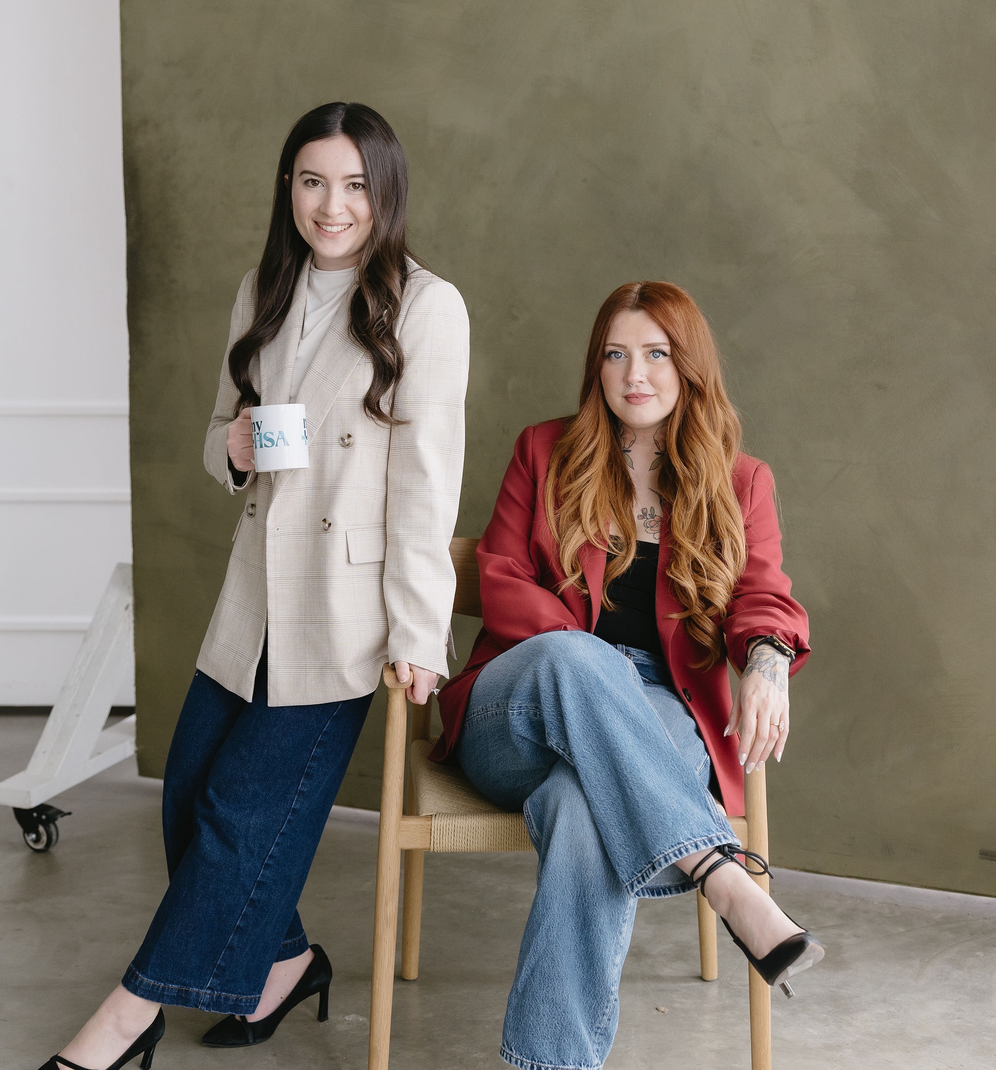 Two women in blazers and jeans posing indoors, one seated and one standing with a coffee mug.