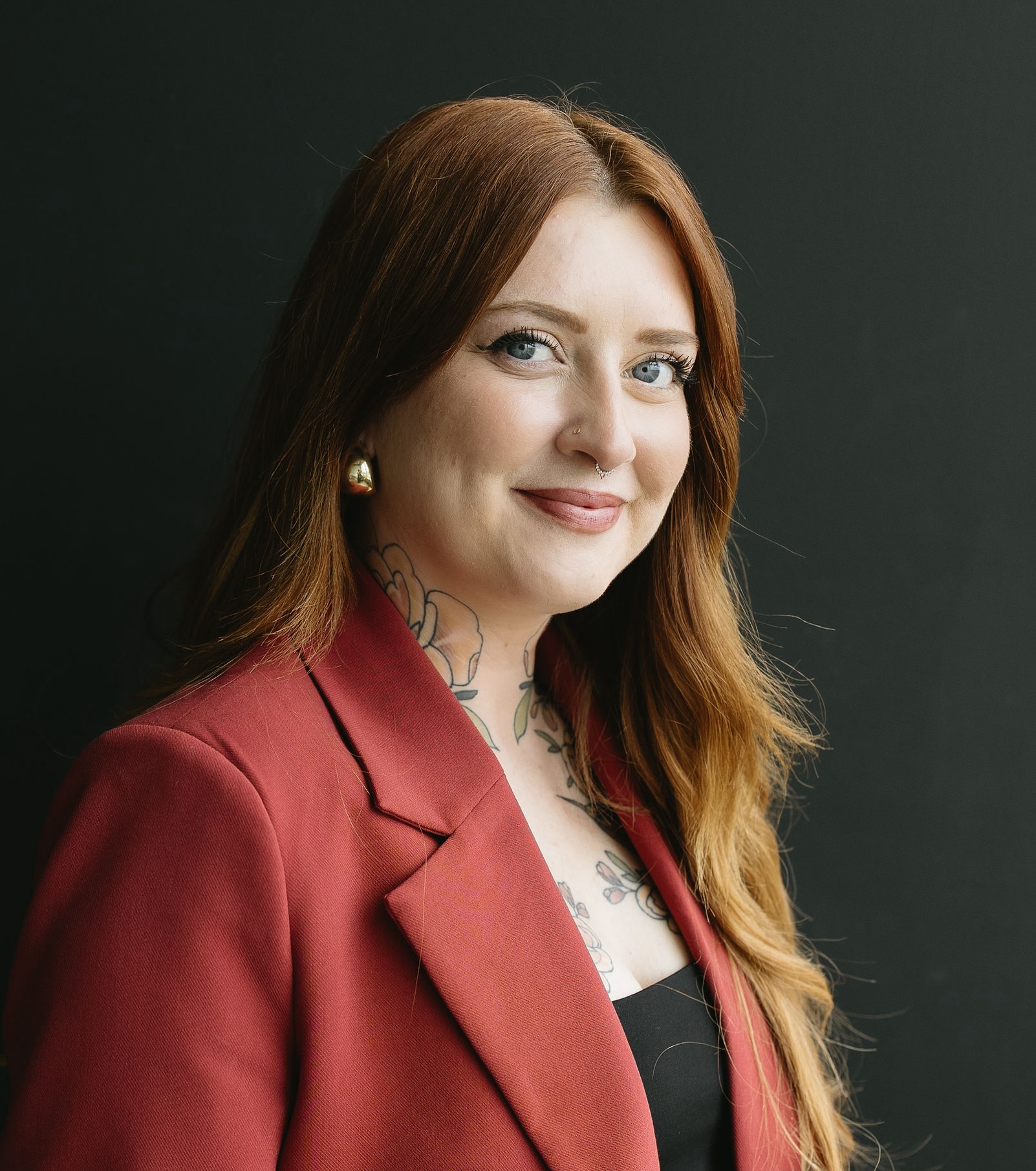 Smiling woman with red hair, nose ring, floral neck tattoo, and red blazer against a dark background.