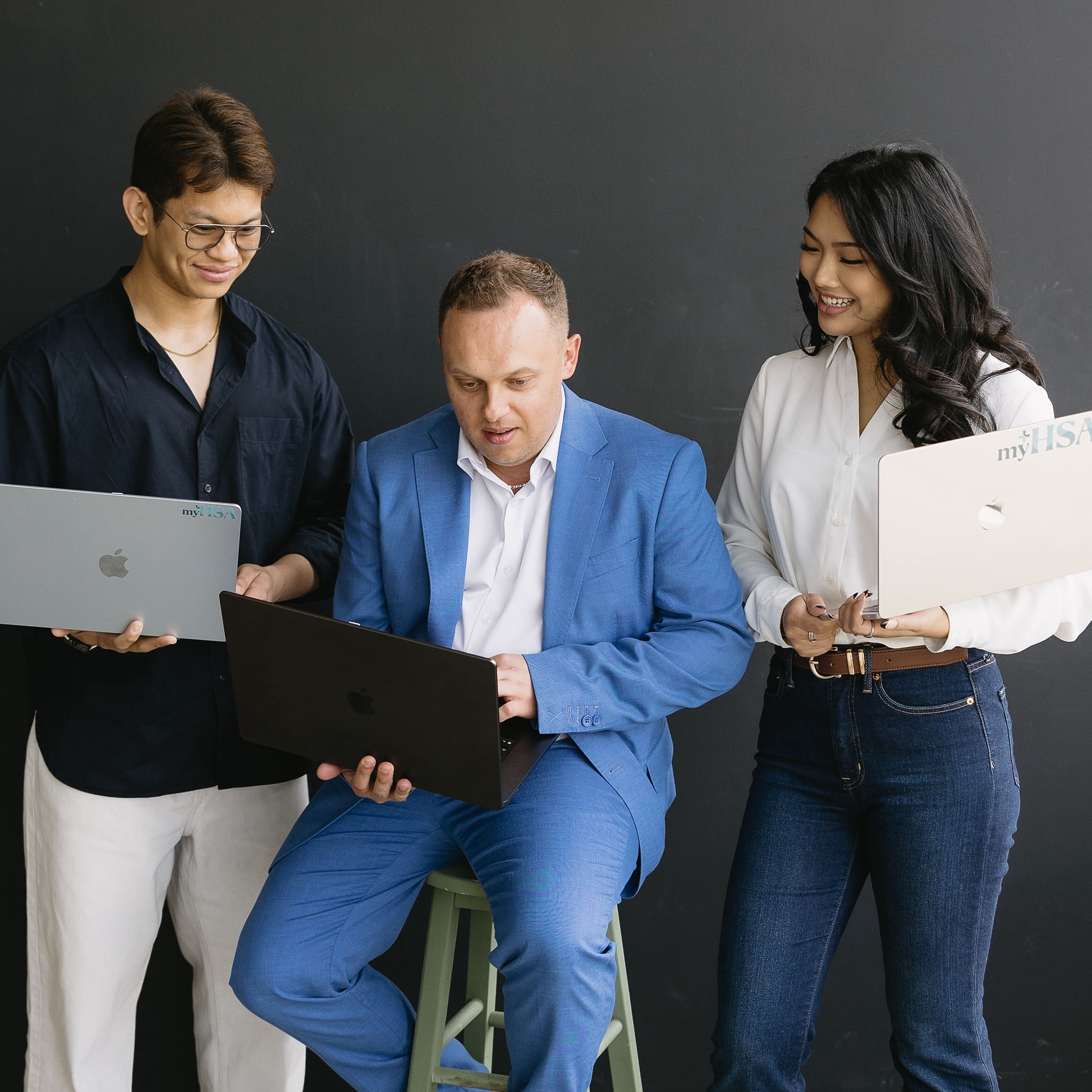 Three people with laptops collaborating against a black background, one seated in a blue suit.