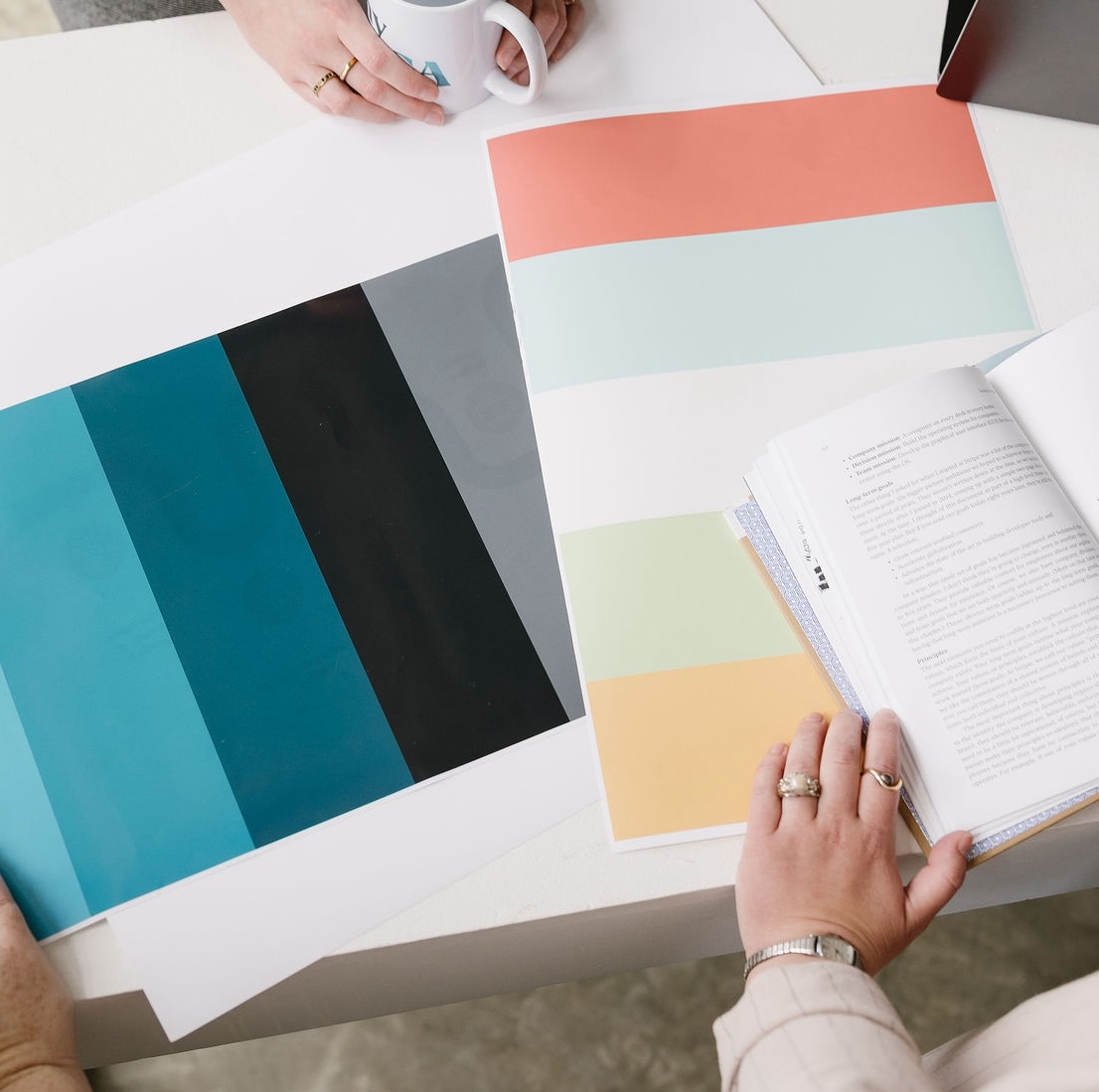 Hands holding a mug and a book near papers with color swatches on a white table.