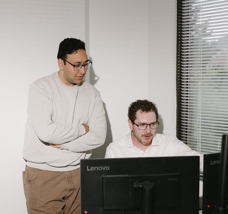 Two men with glasses working together on computers in an office with blinds on the window.