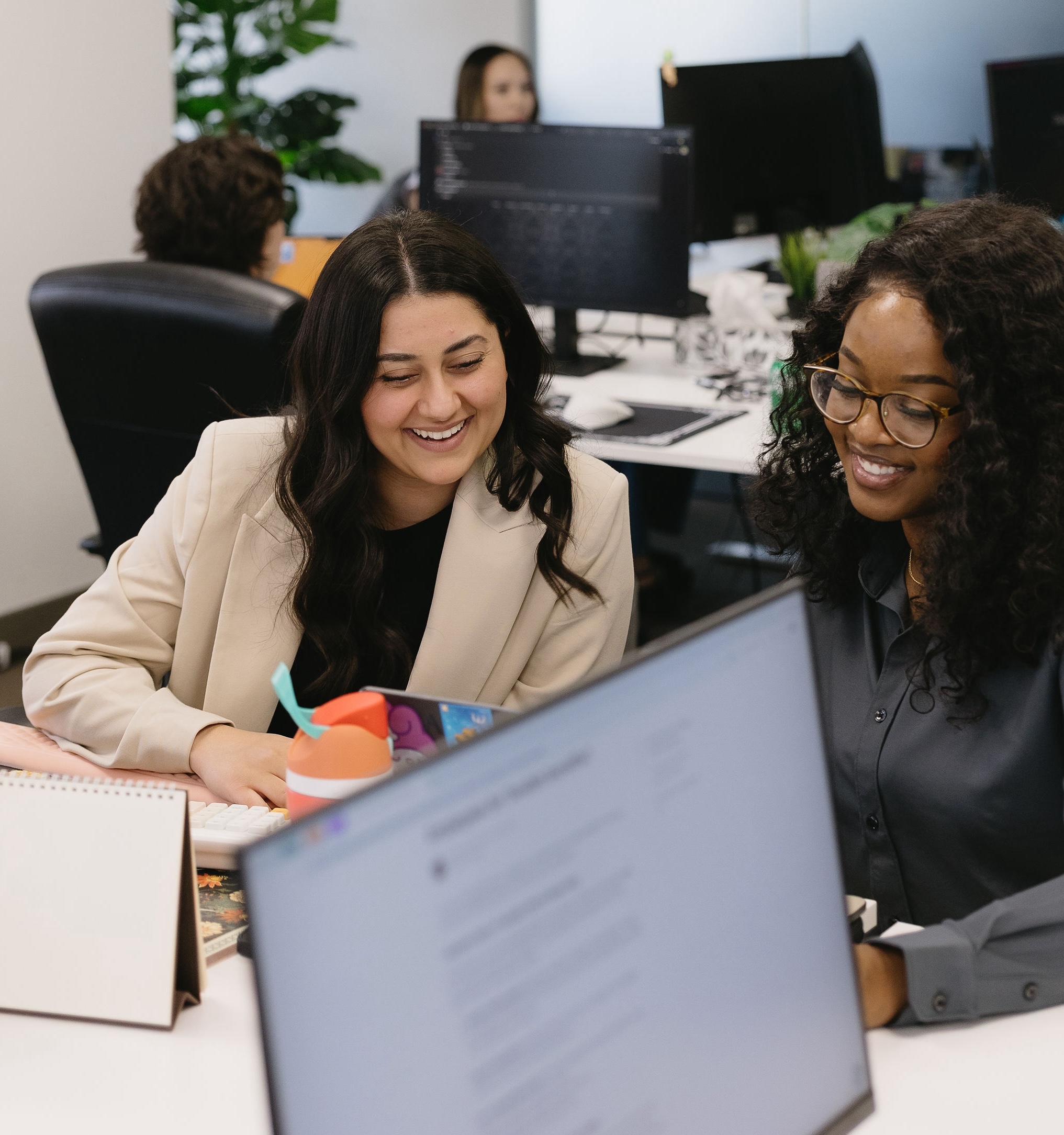 Two women smiling and working together at a desk with computers and office supplies.