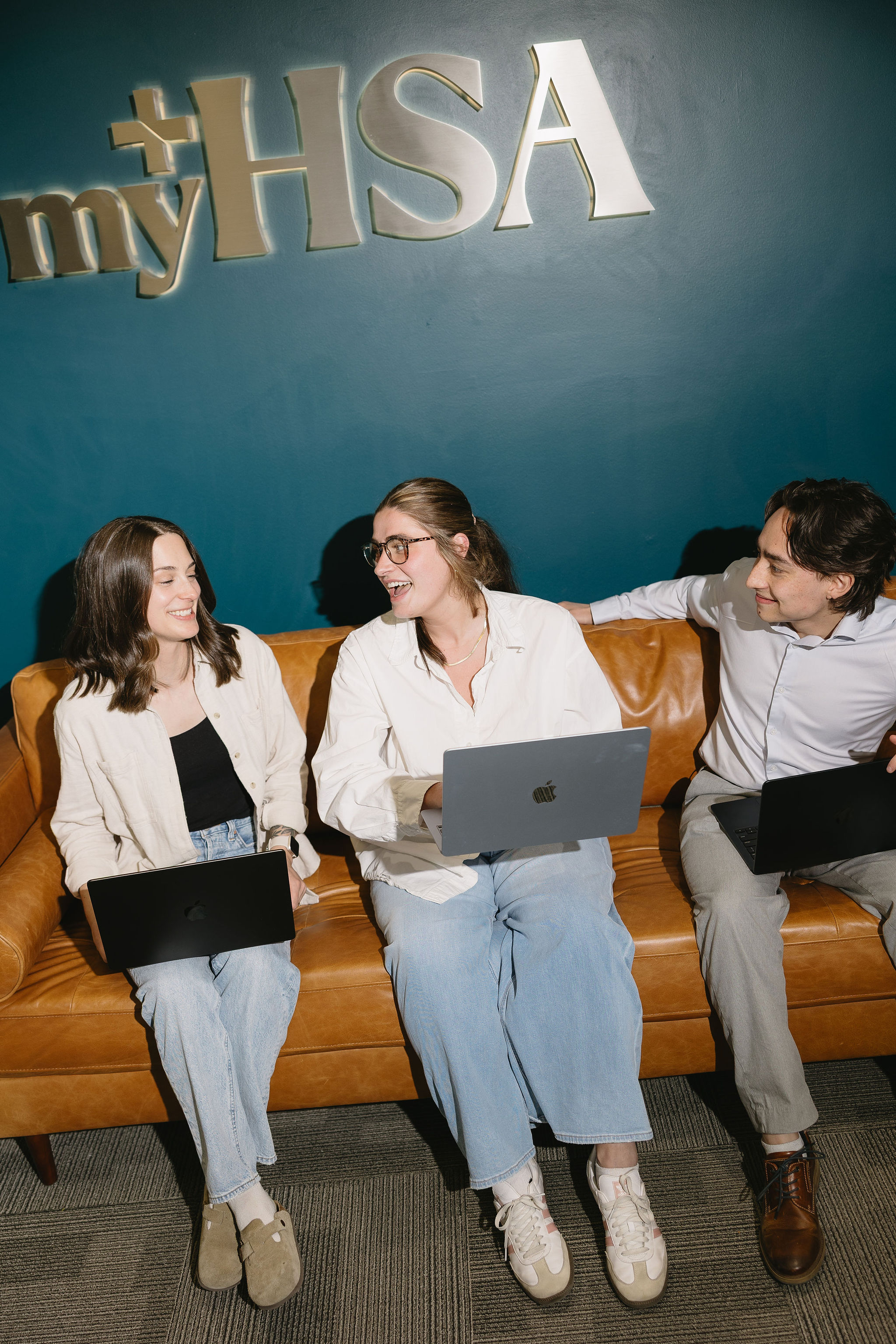 Three young adults on a brown couch with laptops, smiling and talking under a myHSA sign.