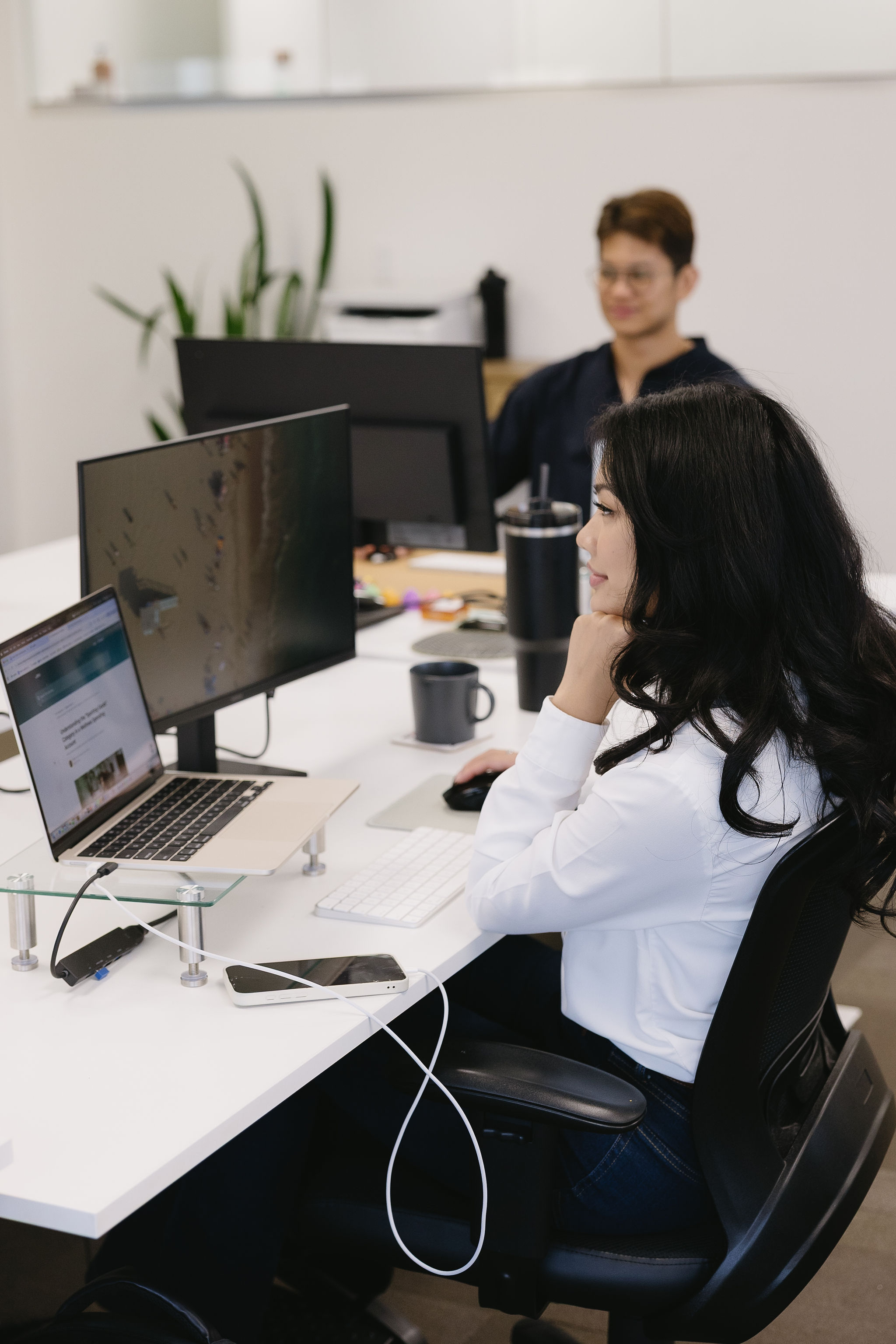 Woman in white shirt working on laptop and desktop at a shared office desk with a man in the background.