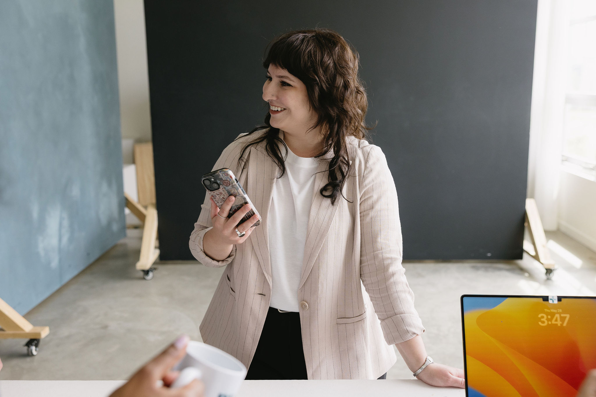 Smiling woman in a beige blazer holding a smartphone standing by a desk with a laptop and coffee mug.
