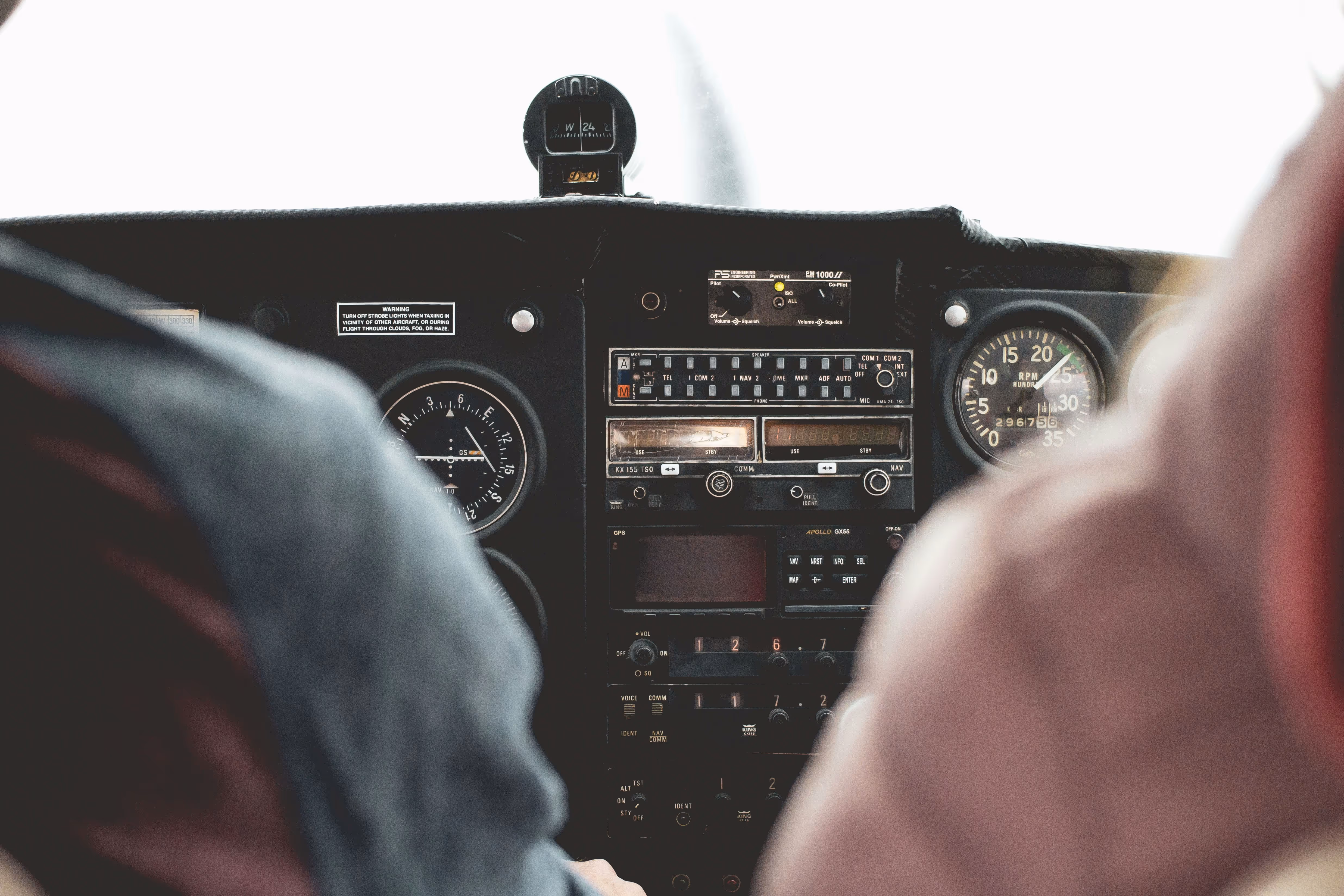 View of an airplane cockpit dashboard with various controls and gauges, seen between two people.