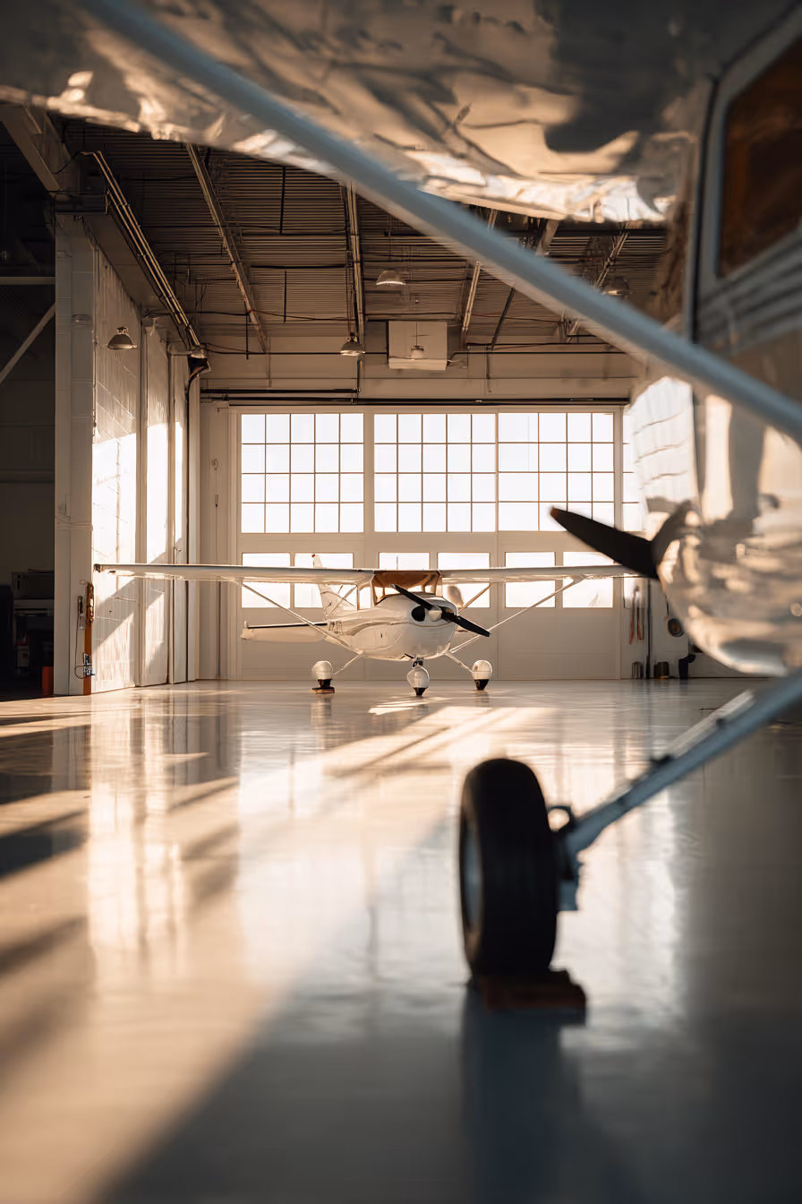 Small white airplane parked inside a bright hangar with sunlight streaming through large windows.