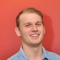 Smiling young man with short light brown hair wearing a blue shirt, standing against a red background.