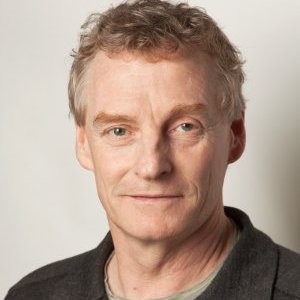 Portrait of a middle-aged man with light curly hair wearing a dark shirt against a plain background.