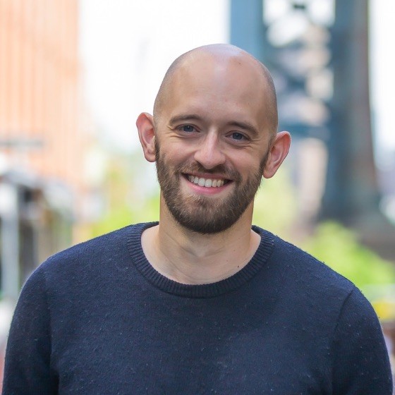 Smiling bald man with a beard wearing a dark sweater, standing outdoors with blurred buildings and greenery in the background.