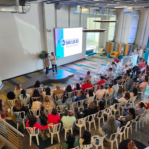 Two presenters speaking on a small stage to an audience seated in white plastic chairs in a modern, bright conference room with a large screen displaying the logo of San Lucas hospital.
