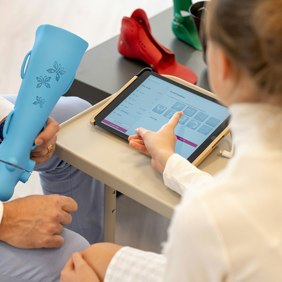 Person holding a blue prosthetic leg while another person uses a tablet showing prosthetic customization options.