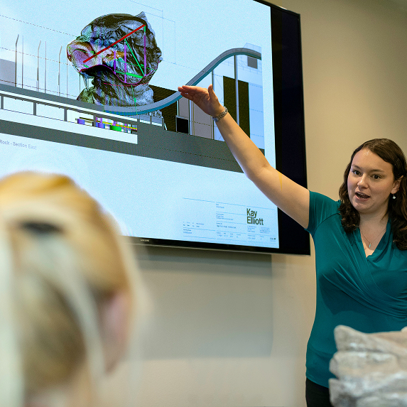 Woman presenting a digital architectural design displayed on a large screen to an audience.