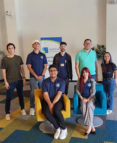 Group of seven diverse young adults indoors, with two seated on colorful modern chairs and a Healthcare Innovation Center sign in the background.