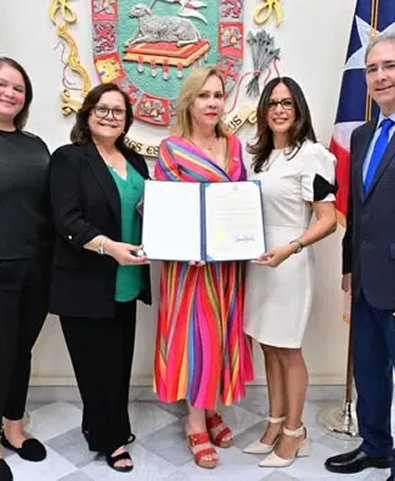 Group of five people standing indoors, with two women holding a certificate in the center, a colorful coat of arms and flags in the background.