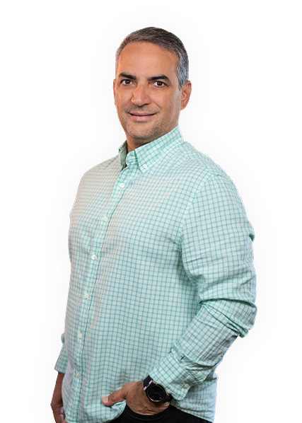 Man wearing a light green checked button-up shirt with a watch on his left wrist, standing and smiling with one hand in his pocket.