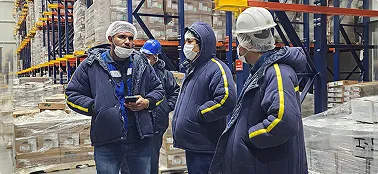 Trabajadores con cascos y mascarillas conversan en un almacén de logística con estanterías llenas de cajas.