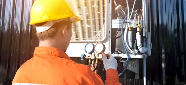 Técnico con casco amarillo revisa manómetros en unidad de aire acondicionado exterior.
