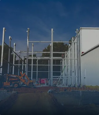 Estructura metálica en construcción junto a un edificio blanco bajo cielo azul.