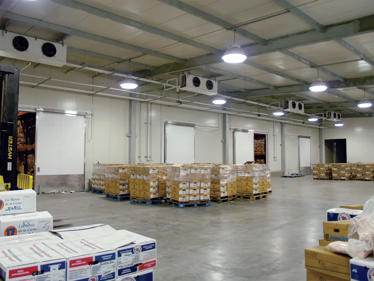 Almacén refrigerado con pallets de cajas apiladas y puerta blanca corrediza abierta.