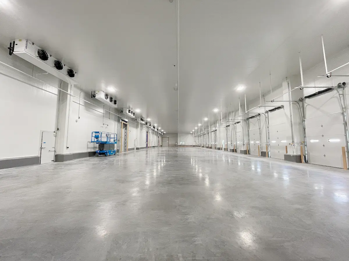 Gran espacio industrial con piso de concreto, luces en techo y puertas grandes a la derecha.