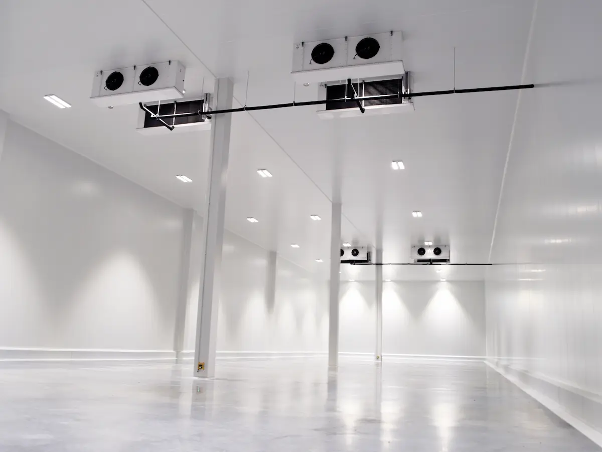 Interior de una cámara frigorífica vacía de gran tamaño con ventiladores en el techo y paredes blancas.