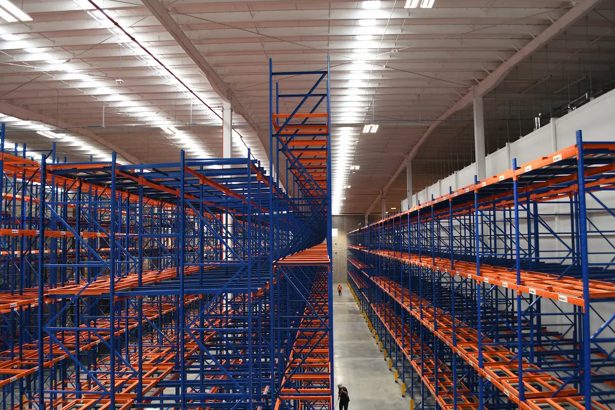Pasillos largos con estanterías de metal azul y naranja vacías en un almacén grande y iluminado.