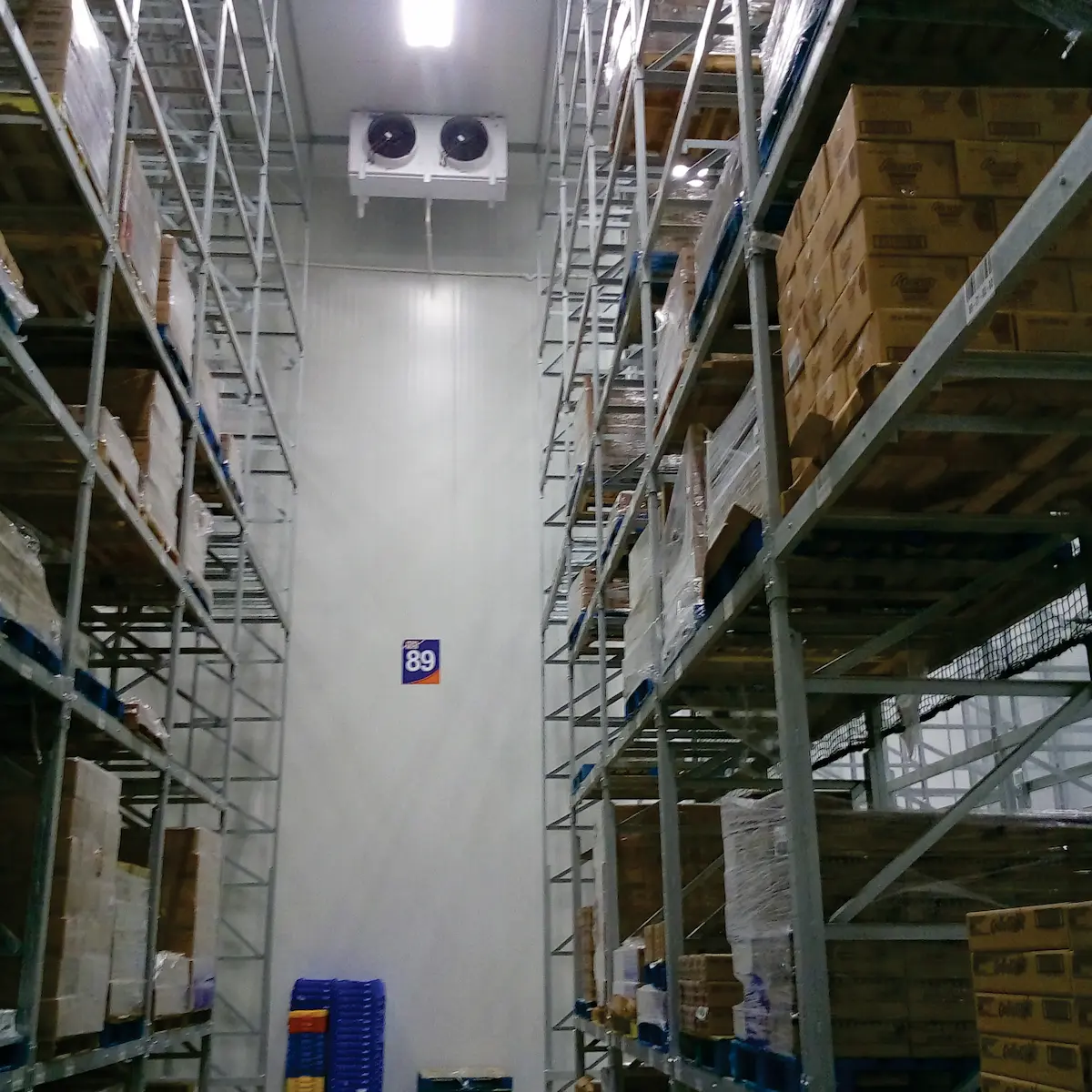 Pasillo de almacen con estanterías metálicas llenas de cajas y ventiladores en la pared.