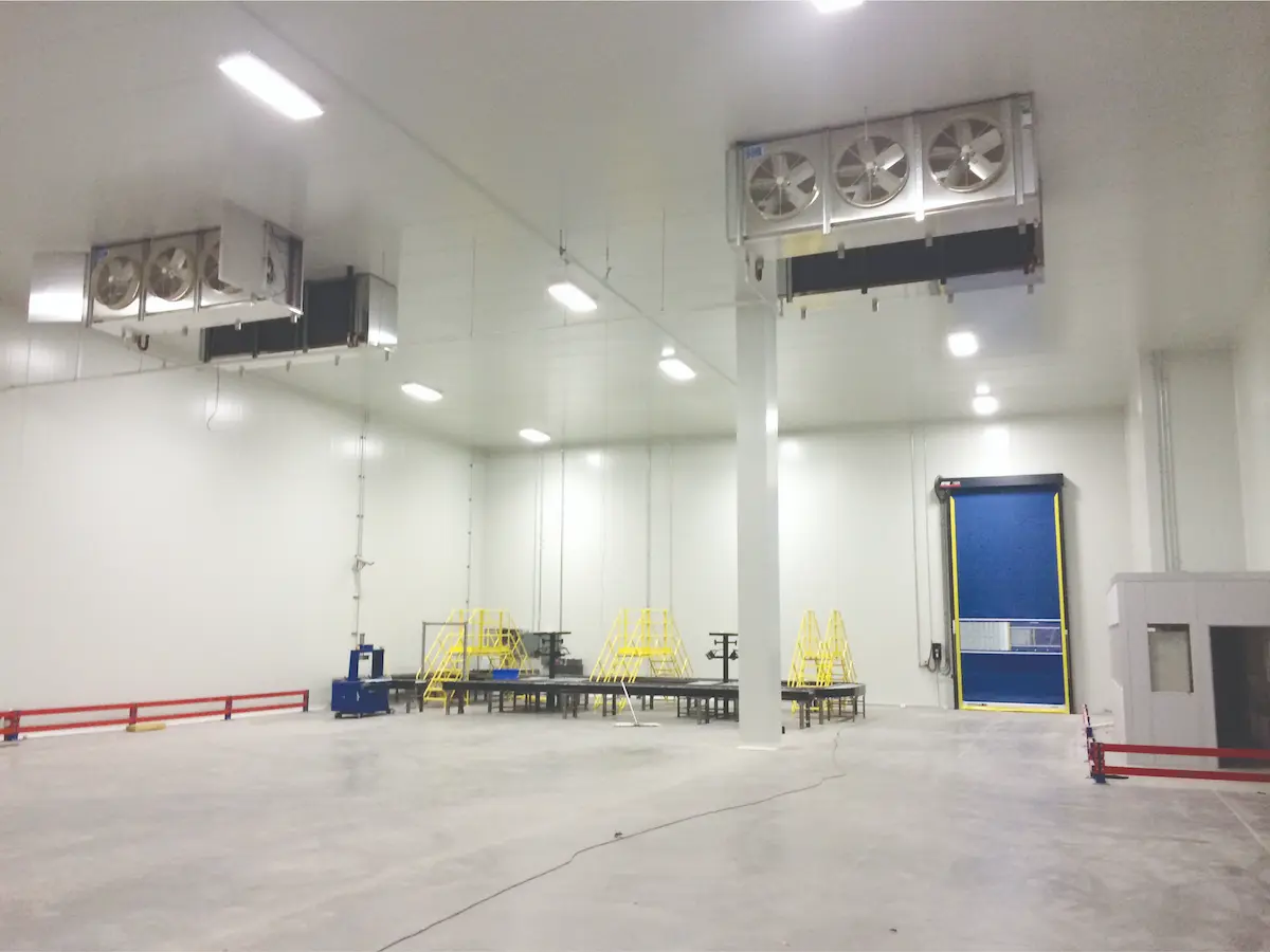 Interior de un almacén industrial vacío con ventiladores en el techo y una puerta azul al fondo.