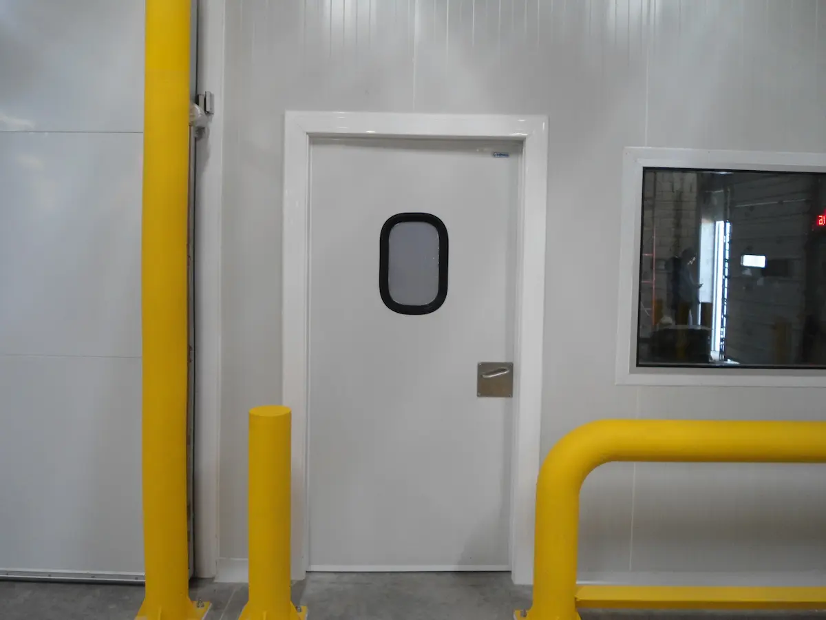 Puerta blanca con ventana ovalada y barandilla amarilla en una instalación industrial.