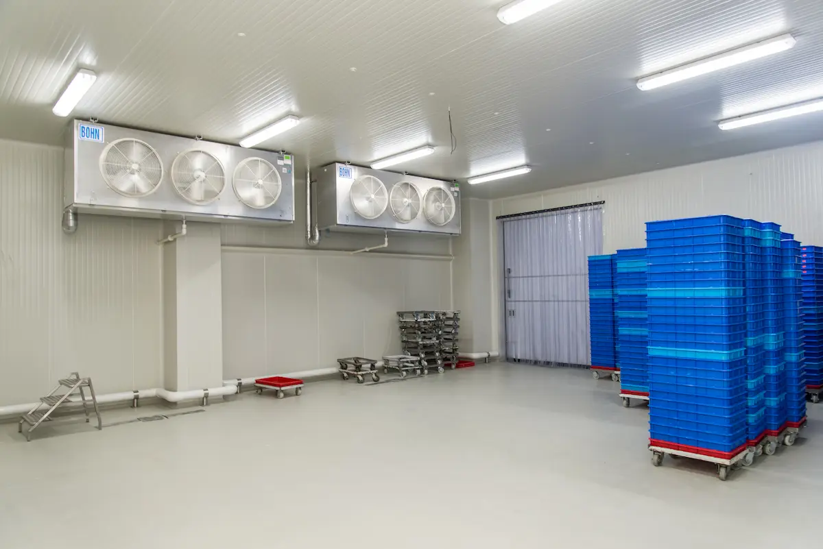 Sala de almacenamiento fría con ventiladores industriales en el techo y pilas de contenedores azules sobre ruedas.